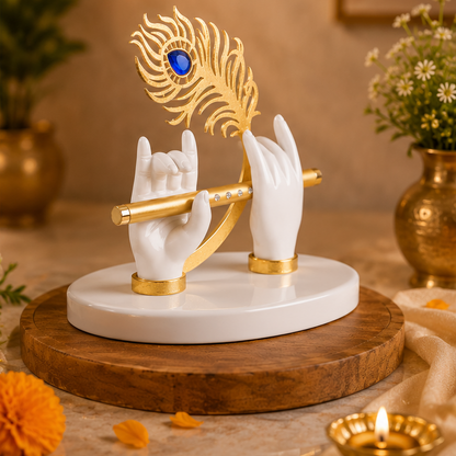 A decorative figurine of two white hands holding a gold flute, with a gold peacock feather featuring a blue stone eye resting above them, displayed on a round white base over a wooden tray in a warm indoor setting.